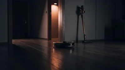 A robot vacuum on a dark wood floor with a cordless stick vacuum against the wall