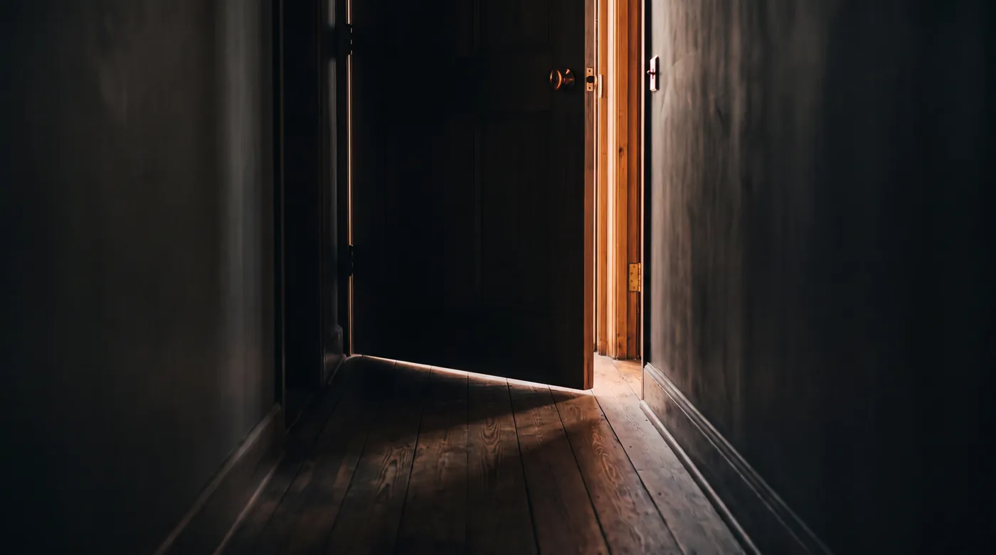A dark hallway with a door left ajar, warm light spilling across the wood floor